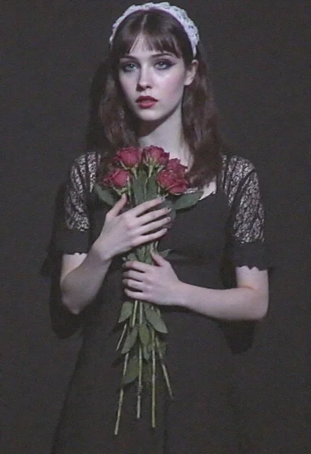 v8s, Young woman, late teens to early 20s, pale complexion, dark straight shoulder-length hair, wearing a vintage-inspired black lace dress, dramatic dark eyeliner, pale foundation, deep red lipstick, white lace headpiece, standing in a mysterious dark setting, holding a bouquet of wilted red roses close to her chest, ethereal goth style, eerie atmosphere,. Amateur quality, Analog distortion, low resolution, Chromatic aberration, tape noise, Grainy texture, Frame jitter, muted colors, overexposed foreground