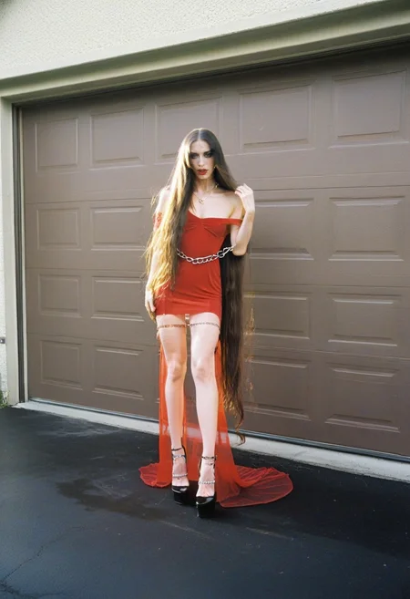 <lora:ModernFable:0.65> woman, surreal, editorial, long hair, red dress, sheer dress, off-shoulder, chains, high heels, black shoes, dramatic makeup, outdoor, bright light, garage door background, suburban setting, strong pose, high contrast, film grain,