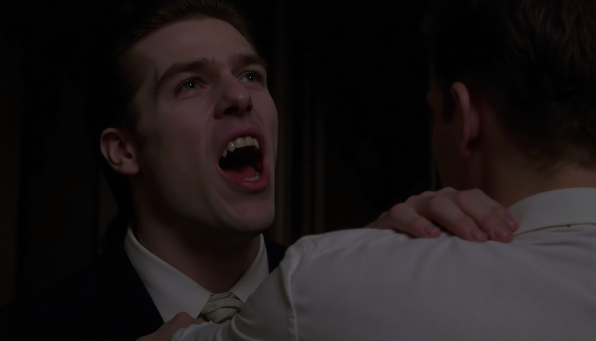 cinematic close-up shot of a vampire man with fangs and very pale skin shown looking over the shoulder of another man, preapring to bite, standing very close, one hand on the back of his neck. The other man is shown from behind, wearing a white shirt. Dark victorian room in the background. At night in soft light. Analogue film, IMAX, photorealistic, very detailed <lora:iwtv_flux_1_0:1>