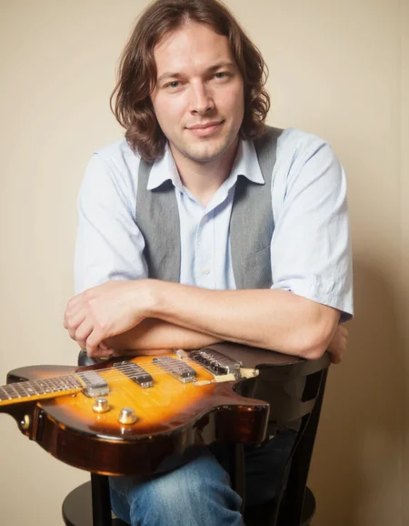 <lora:DAVID_GILMOUR:1>This photograph features a man sitting in a relaxed pose, leaning slightly forward with his arms crossed. He has shoulder-length, dark brown hair that falls naturally around his face. The man is wearing a light blue, short-sleeved button-up shirt with vertical stripes, paired with a grey, sleeveless vest. He is also wearing blue jeans. His expression is calm and slightly smiling.
In front of him, on a black chair, lies an electric guitar with a distinctive sunburst finish, transitioning from a dark brown at the head to a lighter yellow at the body. The guitar's neck is positioned towards the man, with the body resting on the chair. The man's right hand is placed on the guitar, suggesting he may be a musician or enthusiast.
The background consists of a plain, light beige wall, providing a neutral backdrop that highlights the subject. The lighting is soft and even, casting gentle shadows and highlighting the textures of the man's clothing and the guitar. The overall mood of the photograph is casual and intimate, capturing a moment of relaxed enjoyment.