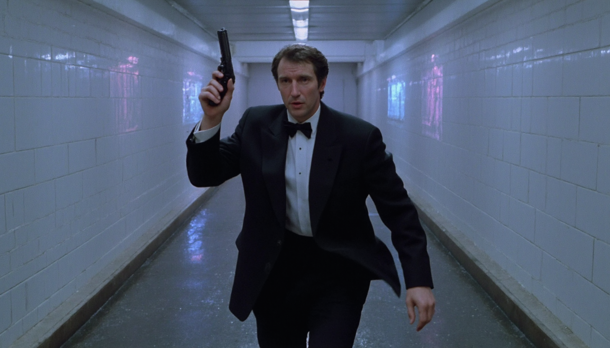 cinematic medium shot of a 40 y.o. agent wearing a tuxedo with a black bow tie, running through a tunnel with white tiled walls and neon lights at the ceiling. The lights reflect in the tiles and on the wet floor. He is holding up a gun. Cold artificial light from above at night. High resolution. 1990s movie <lora:dark_cinematic-000080:1>