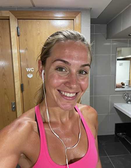 In a medium close-up selfie shot, a young Caucasian woman is in a gym locker room, capturing the moment post-workout. The angle is slightly low and close, showing her full face and upper body as she smiles confidently into the camera. The lighting is artificial, likely from overhead gym lights, casting soft reflections on her sweaty skin. The scene has muted earth tones from the walls and doors, with subtle reflections from mirrors in the background, contributing to a warm, indoor atmosphere. Her skin is visibly shiny and slick with sweat, accentuating her natural skin texture. The light interacts with the moisture on her face and upper chest, creating a sheen that highlights subtle imperfections, such as small pores around her cheeks, forehead, and nose. Faint redness and a slight flush can be seen on her face, likely from the intensity of her workout. The faint texture of small freckles is visible across her upper cheeks and shoulders, and a minor sheen of oil enhances the realism of her skin. Despite the shine, thereâs no excessive smoothness, and the subtle details of natural skin, including small bumps and irregularities, are prominent, especially on the forehead and chin. She is wearing a pink sports bra, which contrasts with her pale skin and is drenched with sweat, clinging to her body. Her blonde hair is pulled back into a practical ponytail, a few damp strands stuck to her forehead and the sides of her face, contributing to the post-exercise look. Her earbuds are tucked into her ears, adding a casual, modern detail to the shot. In the background, the gym locker room features wooden doors marked "WC," tiled walls, and a mirror reflecting part of the room. The lighting is slightly warm, casting soft highlights on the reflective surfaces and her skin. There are no notable technical artifacts or distortions in the image, and while the focus is on the subject, the background adds context without being too distracting. Overall, the lighting interacts naturally with the subject, enhancing the realistic and candid mood of the scene <lora:amateurphoto-v6-forcu:0.8>