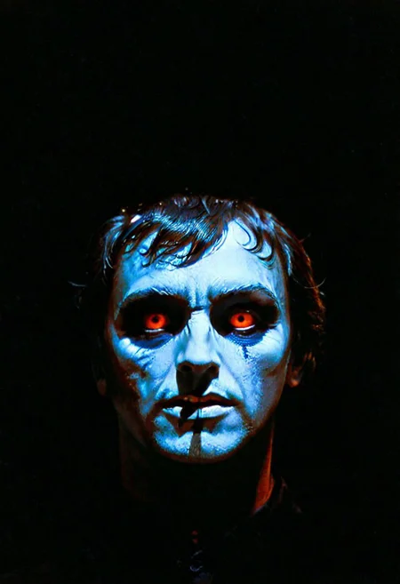 A shadowy silhouette with glowing eyes is standing in a dark room. The darkness of the room and the man's eyes create a striking contrast, drawing attention to the subject. The man is the main focus of the image, and his presence in the dark room adds an element of mystery and intrigue. 1970s horror film