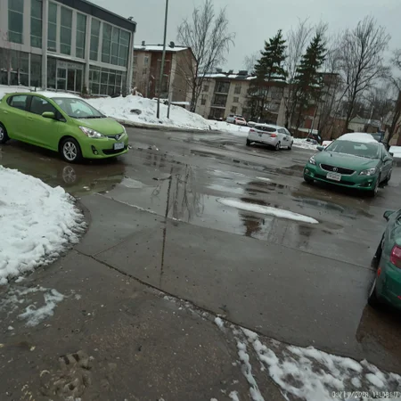 shit_cm_shot, photograph of a snowy parking lot, winter setting, overcast lighting, green compact car in foreground, partially frozen puddle, snow patches on ground, two more cars in background, urban environment, buildings with windows, trees without leaves, slight fisheye lens effect, rule of thirds composition, slight tilt angle, muted colors, cold atmosphere, no people, urban decay,