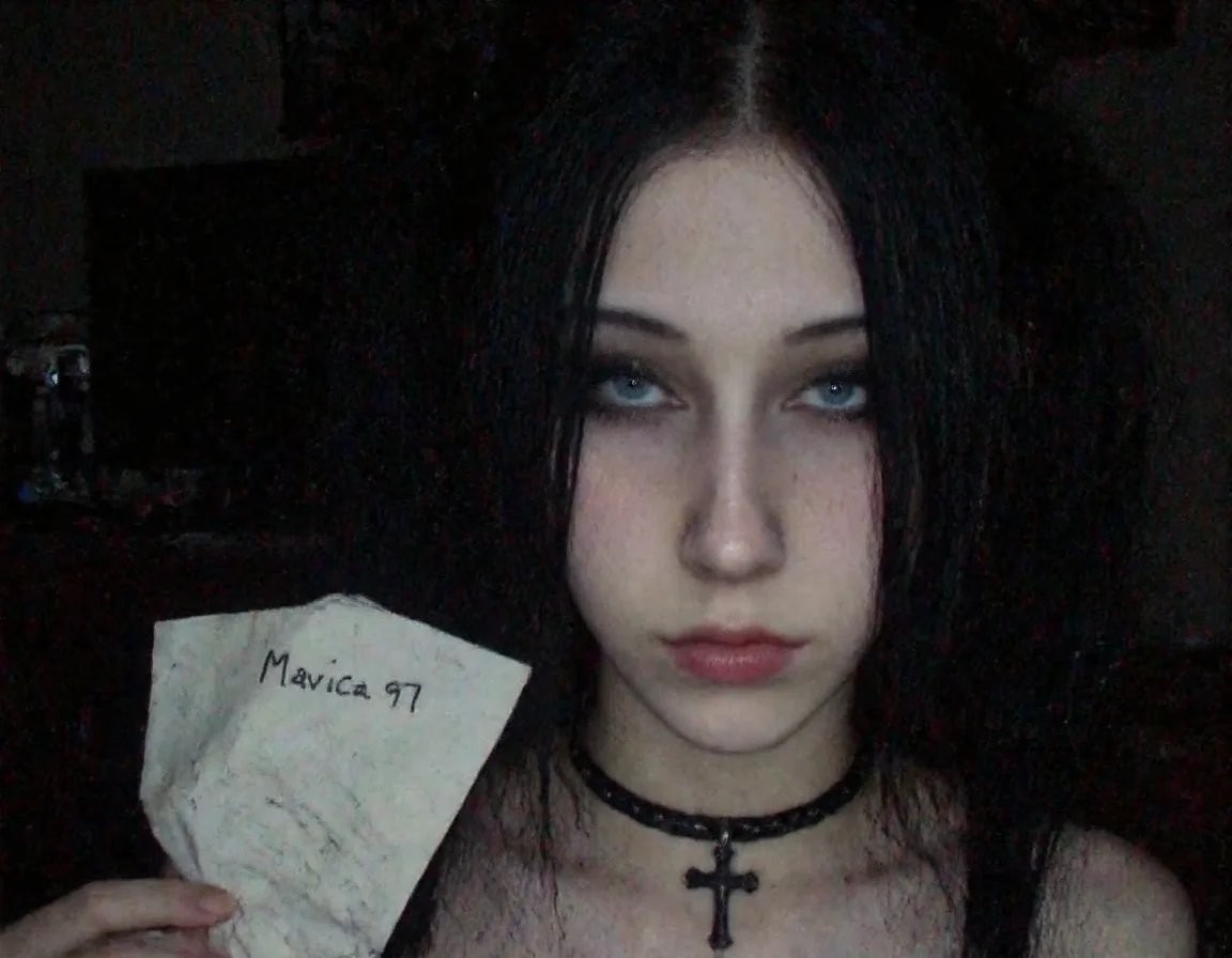 m8vic2, portrait of young woman, early 20s, front view, looking at the viewer, She has light skin and dark hair styled in two pigtails, with bangs covering her forehead, She also has a black choker and a black cross necklace, adding to the gothic aesthetic. Her expression is neutral, right hand near the neck. The background shows a room with furnitures and desk with computer and monitor. Left hand holding a crumpled paper with handwritten text that reads "Mavica 97". dimly lit room, low light condition, moody atmosphere,