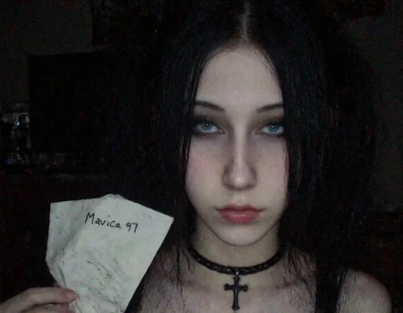 m8vic2, portrait of young woman, early 20s, front view, looking at the viewer, She has light skin and dark hair styled in two pigtails, with bangs covering her forehead, She also has a black choker and a black cross necklace, adding to the gothic aesthetic. Her expression is neutral, right hand near the neck. The background shows a room with furnitures and desk with computer and monitor. Left hand holding a crumpled paper with handwritten text that reads "Mavica 97". dimly lit room, low light condition, moody atmosphere,