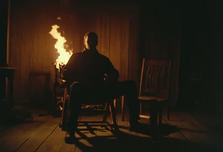 Man sitting on a wooden chair, flames behind him, dark dim lighting, film still, movie scene, shot on 70mm film, Cinematic, shot on kodak vision 3 500t film