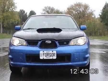 v8s, blue subaru impreza wrx sti, front view, visible headlight details, damaged front bumper, black hood, custom license plate reading "I FORGOR", rainy outdoor setting, trees in the background, overcast sky, reflective wet pavement, horizontal static lines, chroma noise, warped scanlines, "12:43:03" timestamp in bottom right corner