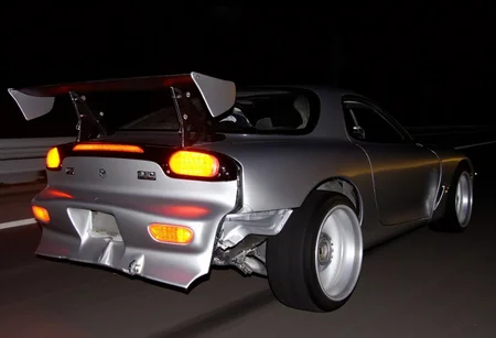 rear three-quarter view of a silver sports car mazda rx7, visibly damaged from a collision. The rear bumper is heavily dented, exposing the internal structure of the taillights. The car appears to be driving on high speed on a dimly lit highway at night, with a broken taillight emitting an orange glow. A black grooved rear wing and aftermarket wheels suggest the car is modified, likely belonging to someone in the drifting or tuner car . The interior is slightly illuminated, showing racing seats with a roll cage. The background reveals a guardrail and a dark, empty road, intensifying the isolation of the scene, amateurish photo, low lighting, in motion, smeared details