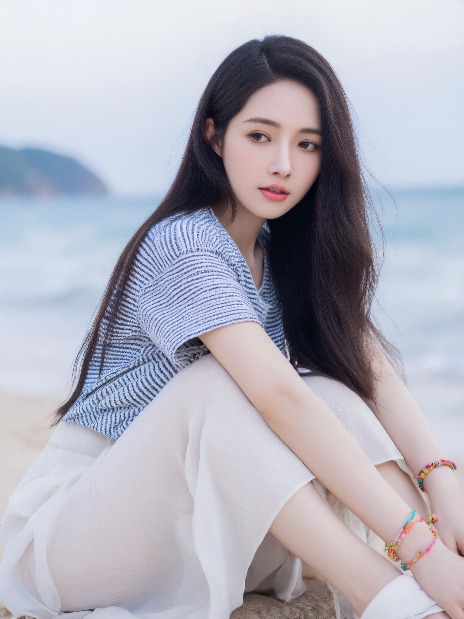 This is a high-resolution close-up photograph of a young Asian woman with long, straight black hair and fair skin. She is sitting with her legs crossed and her right arm resting on her knee. She is wearing a blue and white striped short-sleeved top and a flowing white skirt. Her expression is serene, and she gazes slightly off to the side.

She wears colorful bracelets on her left wrist. Her white sandals are partially visible. The background is softly blurred, showing hints of ocean and sky. The lighting is natural and diffused, creating a calm and gentle mood. The composition follows the rule of thirds, with her face and upper body slightly off-center.