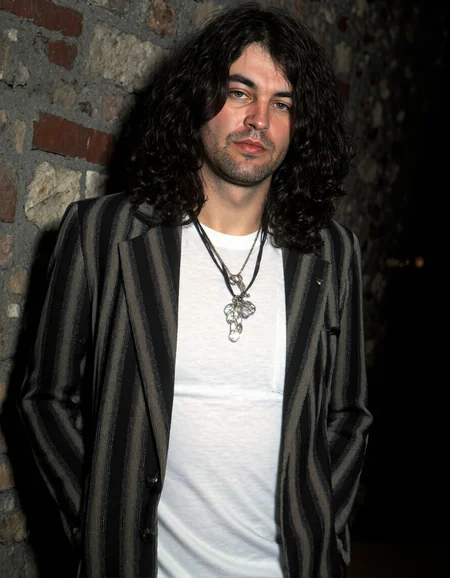 <lora:IAN_GILLAN:1>, This is a photograph of a man with long, dark, wavy hair that falls past his shoulders. He is dressed in a white, long-sleeved shirt and a black and white striped blazer. The blazer has vertical stripes that alternate between black and white. He wears a black tie with a silver pendant in the shape of a butterfly, which hangs prominently from his neck. The man has a serious expression and is slightly smiling. His left hand is casually placed in his pants pocket, while his right arm hangs loosely by his side.
The background of the photograph is a dimly lit, textured wall that appears to be made of stone or brick, with a slightly rough texture. The lighting in the image is soft, casting gentle shadows that enhance the texture of the man's hair and the striped pattern of his blazer. The overall color palette is muted, with the dominant colors being black, white, and shades of grey. The photograph has a candid, natural feel, capturing a moment of relaxed introspection. The image's composition is centered on the man, with the background being slightly blurred, focusing attention on him.
