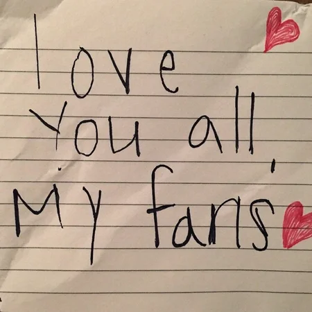 Crumpled piece of lined paper, handwritten message reading "love you all, my fans", written in black ink, neat and legible handwriting, paper background shows visible creases and shadows, natural indoor lighting, close-up angle, focus on text, warm tone lighting, small red hearts interspersed