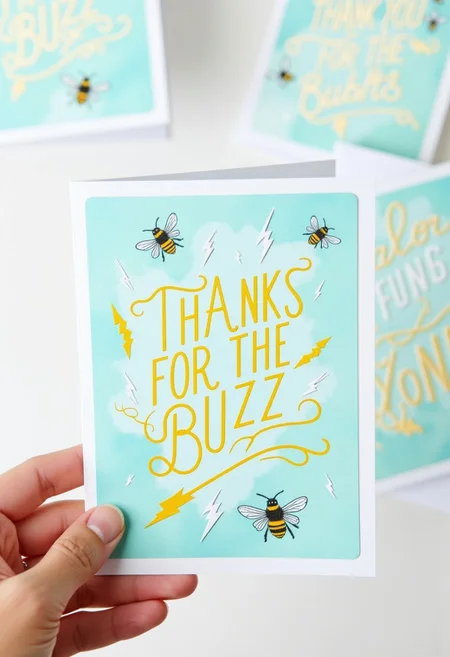 <lora:Greeting_Card_Designer_FLUX:0.7>
The image is a photograph showcasing a beautifully designed thank-you card held by a hand. The card features a beautiful, elegant design with a pastel blue background. The main text, "THANKS FOR THE BUZZ," is written in a bold, elegant script in a yellow color. Surrounding the text are stylized illustrations of lightning bolts and bees. The bees are depicted in shades of black and yellow, with the lightning bolts in varying shades of white and yellow. The illustrations give the card a hand-drawn, watercolor-like appearance with a modern, whimsical touch.
In the background, several other thank-you cards are visible, these cards are also adorned with similar bee and lightning bolt designs, maintaining the consistent aesthetic of the main card.