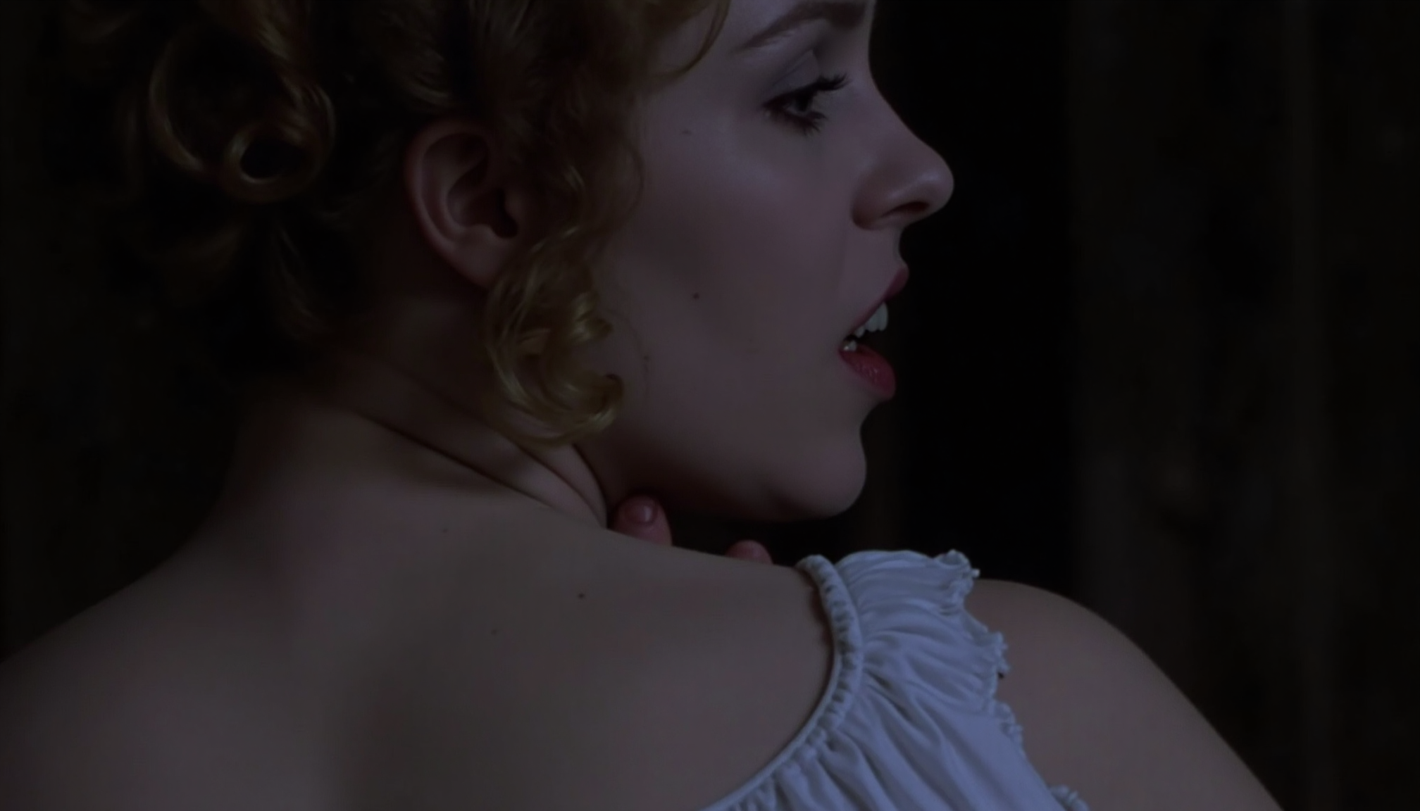 cinematic close-up shot of a vampire woman with fangs and very pale skin shown looking over the shoulder of another woman, preapring to bite, standing very close, one hand on the back of her neck. The other woman is shown from behind, wearing a white nightgown exposing one shoulder. At night in soft light. Analogue film, IMAX, photorealistic, very detailed <lora:iwtv_flux_1_0:1>