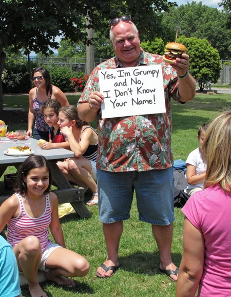 In a wide shot from the front at a family reunion picnic, a grandpa with a massive potbelly and a Hawaiian shirt stands proudly holding a sign in one hand and a burger in the other. Heâs got a mischievous grin, sunglasses perched on his bald head, and the sign reads: "Yes, I'm Grumpy, and No, I Donât Know Your Name!" Around him, kids are running wild, family members are either laughing or giving him side-eye, and grandma is shaking her head with an exasperated smile. Behind him, the picnic table is overflowing with food, and someone is trying to get the grill under control. But grandpa? Heâs living his best, grumpy life in the middle of the chaos <lora:amateurphoto-v6-forcu:0.8>