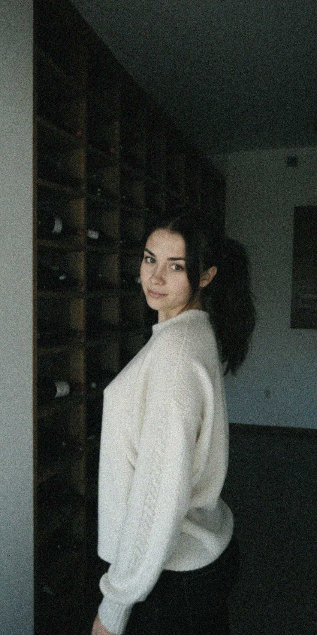 (raw, light leak, film grain, jpeg artifacts, low res, low quality:1.3), shot on GoPro Hero, , 20 year old woman, white woman, tanned skin, slender body, ponytail hair, jet black hair color, cream chunky knit oversized sweater, round neck, worn with black skinny jeans, small home wine cellar or closet conversion, insulated walls, simple wooden wine racks holding numerous bottles horizontally, temperature control unit possibly visible, low consistent lighting, Morning