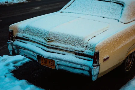 8n8log, Vintage cadillac deville 1970, rear portion visible, completely covered in fresh snow, chrome bumper partially visible, yellow license plate partially showing, parked outdoors, sunset setting, dark asphalt surface with snow patches, cold winter environment, bluish tint from ambient lighting, underexposed image, snow accumulated on trunk and rear window, chrome trim details visible beneath snow layer, low light conditions creating dark shadows around vehicle