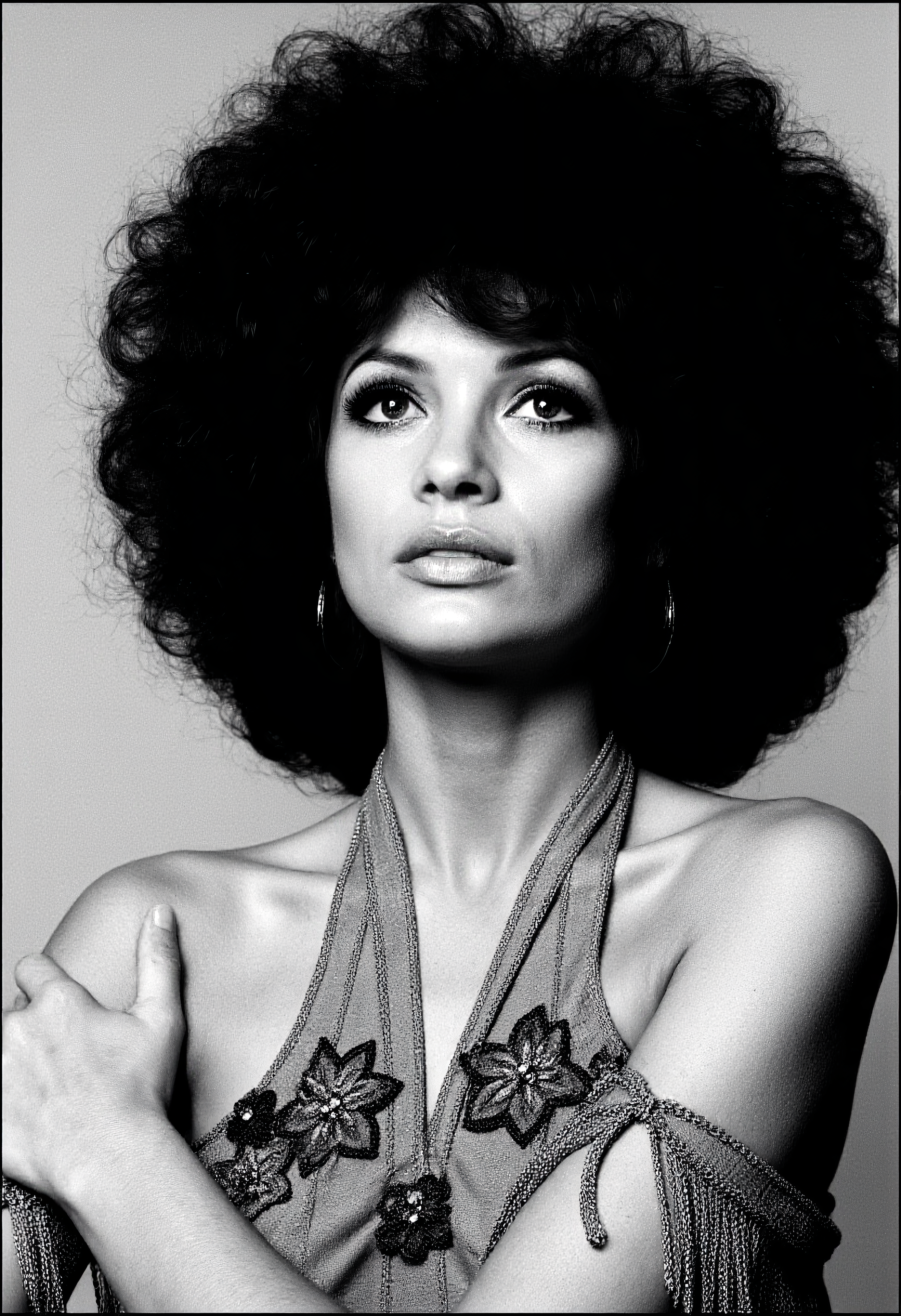 A high-contrast photo of a glamorous woman with a voluminous afro hairstyle, captured in a striking black and white portrait. She is elegantly posing for a picture, her expressive eyes adorned with dramatic eyelashes, gazing upward with confidence and grace. The composition includes intricate floral embroidery and fringed details on her draped outfit, evoking a 1960s aesthetic. The lighting accentuates her radiant features, and the style is heavily inspired by the iconic photography of david-bailey, emphasizing bold contrasts and timeless elegance. <lora:david-bailey-flux1-dev-v1:1>