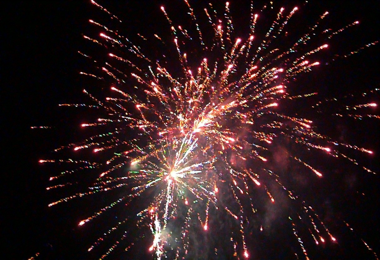 m8vic2, Bright colorful fireworks display, multiple bursts with red, orange, white, and green sparks, night sky background, dense smoke trails, vivid streaks of light radiating outward, outdoor setting, dark atmosphere, overexposed light from fireworks bursts, no visible text or signage, dynamic view upward., 