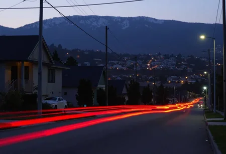 Suburban street scene at dusk, dark houses with muted colors, illuminated by streetlights. Long exposure creates red streaks of car tail lights curving along the road, leading towards the horizon. Overhead, power lines cross the sky, with a distant freeway visible on an elevated section. Background shows a hillside dotted with more homes, shrouded in a light mist, evoking a quiet, sleepy neighborhood. Image has bokeh, soft focus in the background, long exposure, professional quality., 