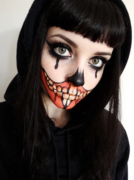 s2ms8ng, girl, early teens, striking green eyes, dark smokey eye makeup, long lashes, black tears painted down from lower eyelids, small freckles across nose and cheeks, intricate face paint resembling stylized skeletal teeth in vibrant red, orange, and black hues covering her lips and extending across her cheeks, a vertical red and black painted stripe running down from the center of her lower lip, straight black hair with blunt bangs framing her face, wearing a black hooded sweatshirt with the hood up,  neutral expression with a direct gaze at the camera, Dreamy soft focus