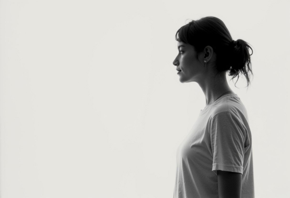 an aesthetic, minimalist depiction of a female profile in side view. she wears a t-shirt. The focus is on the soft contours and calm, monochromatic tones. The scene feels mystical and dreamy, almost as if viewed through a delicate mist, with gentle light accentuating the silhouette. The background is diffuse and creamy white, drawing attention to the elegant simplicity of the figure.