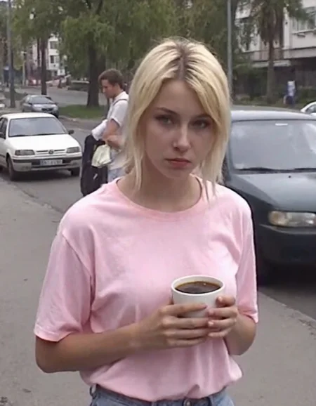 v8s, young woman, late 20s casually dressed in an oversized pink T-shirt, outdoors, her gaze directed to the side, sad expression, hold cup of coffee, her blonde hair loosely tied back. Her face partially shadowed, minimal makeup visible. she is standing on the street near soviet-era storey buildings, trees, parked cars near. Dull, overcast lighting. Amateur quality, Analog distortion, low resolution, Chromatic aberration, tape noise, Grainy texture, Frame jitter, muted colors, overexposed foreground