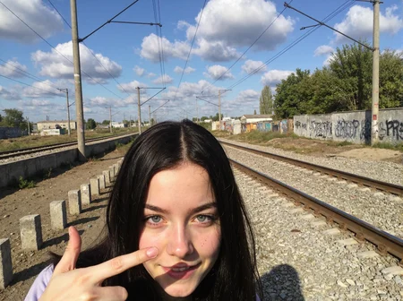 l3n0v0. closeup portrait of teenager girl, blue eyes, black long hair, she is showing piece sign, relaxed expression. Railroad tracks stretching into distance, parallel electrified lines overhead, wooden utility poles with power cables, concrete barriers with graffiti on both sides, dirt path alongside tracks, green trees visible, industrial buildings in far distance, bright blue sky with white puffy clouds, late afternoon natural lighting, urban industrial setting, weathered concrete posts in foreground, gravel railway bed, abandoned industrial appearance, natural spring daylight, wide-angle perspective. amateur quality, candid style
