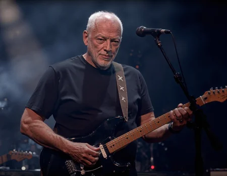 <lora:DAVID_GILMOUR:1>, The image is a high-resolution photograph capturing a live performance by an older man playing an electric guitar. The man, who has short white hair and a beard, is centrally positioned and appears to be deeply engaged in playing. He is wearing a plain black t-shirt and has a brown leather strap across his shoulder, which is reminiscent of a guitar strap. The strap has decorative stitching that adds a textured detail to his attire. The man is holding a black electric guitar with white pickguards and silver hardware, including the tuning pegs and bridge.,
The background is dark and slightly blurry, suggesting a concert setting with stage lights illuminating the performer. The stage lights create a dramatic contrast, casting a soft glow around the man and highlighting his figure. A microphone stand is visible on the right side of the image, indicating that he is likely singing or speaking into the microphone. The overall ambiance of the photograph is energetic and intense, capturing the essence of a live music performance. The textures in the image range from the smooth, polished surface of the guitar to the rougher texture of the man's beard and the leather strap.