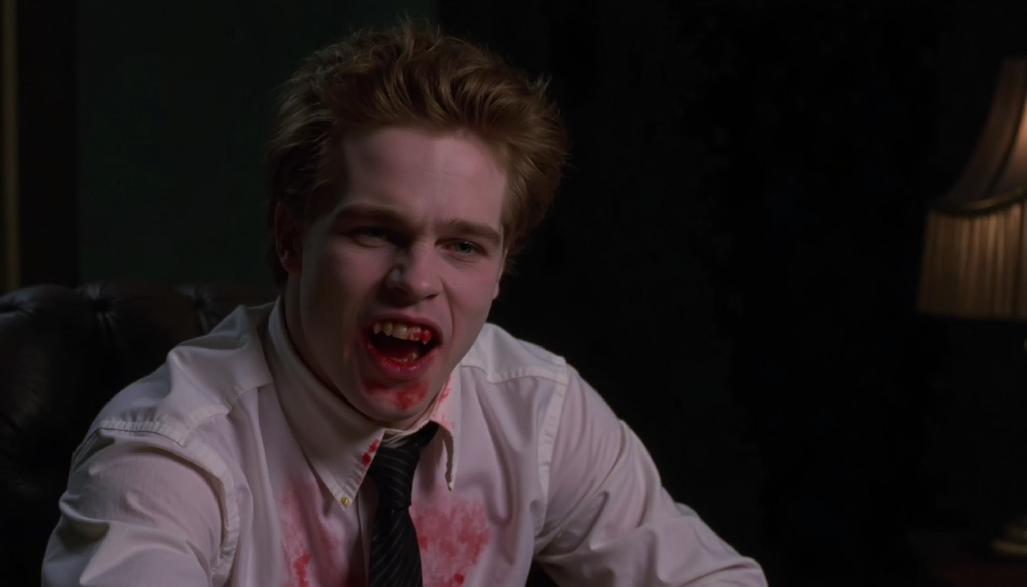 cinematic close-up shot of a young vampire business man with fangs sitting on a desk, looking intensely at the viewer, very pale skin, mouth open showing fangs, blood around mouth, blood on white shirt, loosened tie, dark suit, messy hair hanging over face, lit from one side by a desk lamp with cold light, shadow on the other side, strong contrast between pale skin and blood, analogue film, IMAX, photorealistic, very detailed <lora:iwtv_flux_1_0:1>