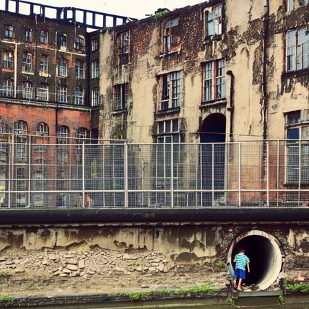 long shot, ruins, under building, wall corner, 1boy behind metal industrial fence, metal grid, looking inside hole, tunnel, window frame, cracked, mud, urban, moody, canalside, building construction, oversaturated