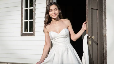 Candid photography of a close-up view of a woman stepping out of a country chapel in a stunning white wedding dress, with the image focused on the upper body. Her dress is beautifully detailed, with intricate lace patterns and a flowing design. She holds the skirt gently with one hand, and her expression is radiant, filled with joy and love. The soft sunlight highlights the details of her dress and illuminates her face, adding a glowing effect. The full view of her face and the upper part of her dress are visible, with the background showing part of the charming wooden chapel. <lora:Sherry-00:1><lora:SelestaFlux1:1>