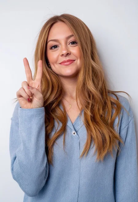 The image is a high-resolution photograph of an amateur actress wearing a long-sleeved top in front of a plain white background. She is casually giving the peace sign. The image captures her from the shoulders up, providing an intimate, close-up view of her face and upper body. She has a fit womanly physique and is slim and slender with medium-sized breasts. The photo is impressively detailed.