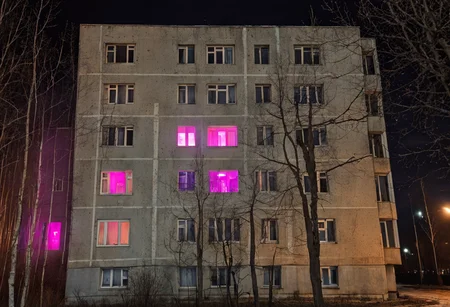 A night-time view of a monolithic, Soviet-style residential building with stark facades lit by harsh artificial light, creating a melancholic atmosphere. Bare trees in the foreground obscure the lower levels, enhancing the eerie mood. Several windows emit a warm glow, but two particular windows stand out with an ominous purple light, adding a touch of the uncanny against the otherwise drab exterior. The building's uniform pattern and the scarcity of visible life convey a sense of desolation typical of the Russian suburbs. The scene evokes feelings of isolation and bleakness. architecture, night, Russian suburbs, Soviet building, eerie, melancholic, isolation, bare trees, purple light, apartment building, desolation, film grain professional 35 mm photo, tumblr <lora:dark slav suburbs_v0.0.1-15:0.75>