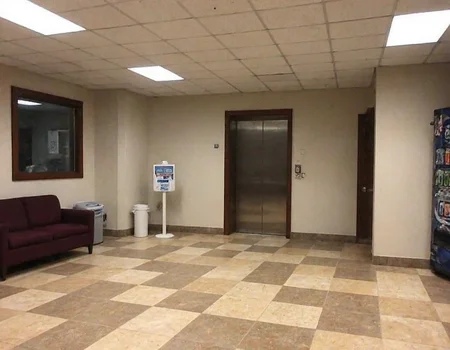 m8vic2, Quiet indoor setting, beige and brown-toned tiles on the floor, elevator door on the right, vending machine partially visible on the far right, maroon couch in the lower left corner, hand sanitizing station against the left wall, large trash bin next to the sanitizing station, beige walls with darker ceiling borders, fluorescent ceiling lights providing illumination, no natural light, hallway environment, appears empty and clean, commercial building or office space, standard office ceiling tiles, mixed lighting conditions with artificial overhead lights.,