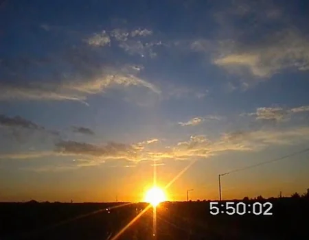v8s, sunrise over horizon, vivid blue and orange gradient sky, scattered clouds, bright sun with lens flare, timestamp "5:50:02", silhouette of landscape and road visible, serene outdoor setting.