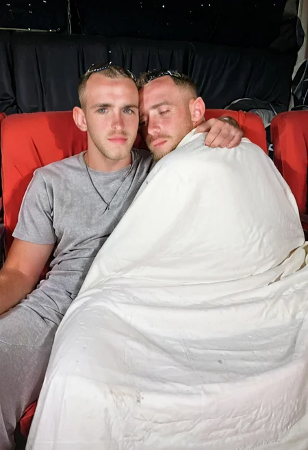 A man sleeping in a movie theater bench seat church pew with a blanket snuggled up next to another male. They are both covered up in a blanket, snuggled together, head on shoulder,