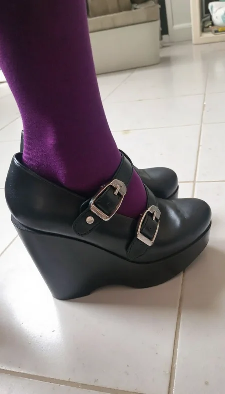 Platform shoes, black leather, two straps with silver buckles, worn over purple and black striped socks. Shoes resting on a white tiled floor, minimal background, focus on footwear, slight vignetting around edges, high contrast, overall clear image quality.