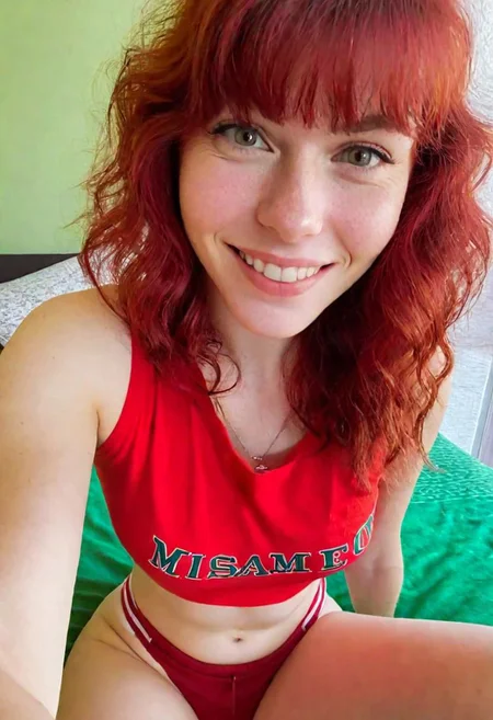 A full-body "Selfie taken with the front camera of an iPhone 15 in a close-up shot." female figure, a smiling and sexy redhead with bangs and wavy hair, dressed in a red and red t-shirt, and green shorts, sitting on a bed, looking at the camera, you can see her perfect breasts. , captured with a blurred background in an elegant hotel room. in leonard style, there is a natural light source illuminating the interior from a window, the background is blurred, the second part in hyper realistic style is made in a special test, by alex1shved, very realistic, rosy skin, beautiful lips and fleshy, smile, feeling of lightness and joy, hyperrealism, highly elaborate skin, direct gaze, by alex1shved