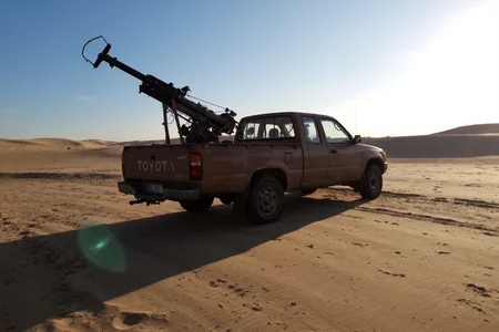 s2ms8ng.  Brown rusty 1990s Toyota Hilux pickup in the desert with a 155mm howitzer mounted in the bed and turned upward, bright sun casts lens flares, overexposed highlights, harsh shadows stretching across the sand.