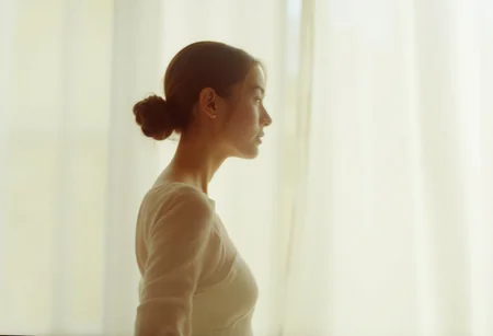 an aesthetic, minimalist depiction of a female profile in side view. The focus is on the soft contours and calm, monochromatic tones. The scene feels mystical and dreamy, almost as if viewed through a delicate mist, with gentle light accentuating the silhouette. The background is diffuse and creamy white, drawing attention to the elegant simplicity of the figure.
