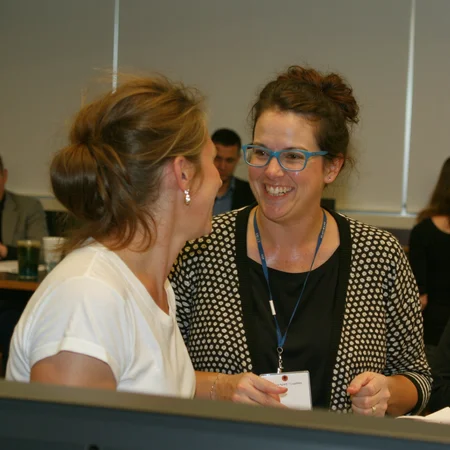 photo of two womenn talking and hugging while working at a busy place. flikr photo from 2012. three people working in the background.