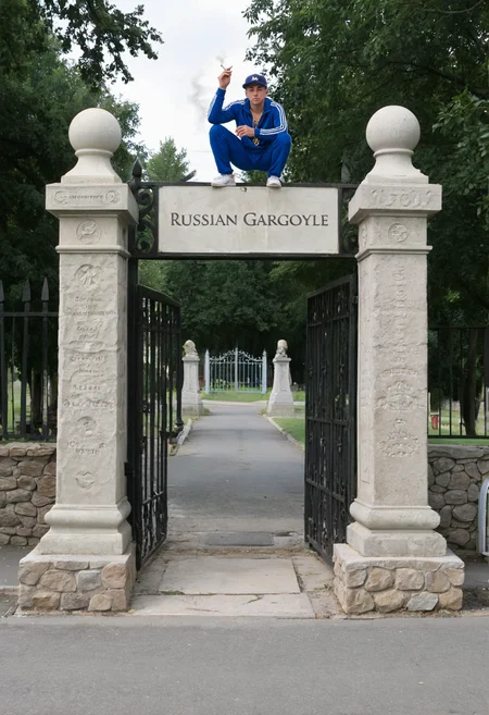 Photo of a cemetery entrance with a large iron gate flanked by two old stone pillars connecting to a stone wall. A man in a blue tracksuit is squatting on top of one of the stone pillars next to the gate. The man is wearing a cap and smoking a cigarette. The background shows a cemetery behind the gate. A text caption at the bottom reads: "Russian Gargoyle" <lora:xl_slavpunk_russia_aesthetic_v1>