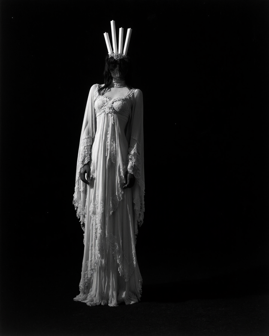 Kaos, The image is a black and white photograph of a woman standing against a stark black background, creating a dramatic, high-contrast scene. The woman is dressed in an elaborate, Gothic-inspired costume that resembles a Victorian-era mourning outfit, but with a surreal, theatrical twist. She is draped in a flowing, sheer, white fabric that cascades down to the floor, creating a ghostly, ethereal effect. The fabric is adorned with intricate lace patterns and delicate fringes, adding to the ethereal quality of her attire.
Her head is adorned with a headdress that features five tall, white candles arranged in a line above her head, creating a striking silhouette against the black background.