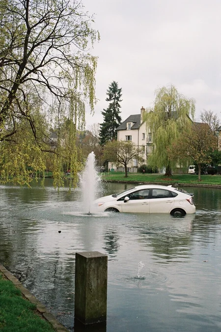 8n8log, Park pond or small lake, calm water surface with slight ripples, weeping willow tree with partially bare branches and light yellow-green leaves, stone fountain or water feature in center of pond creating vertical water spray, white toyota prius is drowning in the pond, white or light-colored building visible in background through trees, additional trees with various foliage visible along shoreline, cloudy or overcast natural lighting, late afternoon or early evening time, residential area visible beyond water, spring or early autumn season based on tree foliage, grassy areas visible along pond edges, muted color palette with blue-grey water tones and pale yellow-green foliage, peaceful suburban or urban park setting, slightly faded or vintage photographic quality. film photography aesthetic, film grain effect prominent throughout image, high contrast lighting creating dramatic shadows, grainy film-like texture