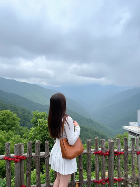 The image is a photograph capturing a serene mountainous landscape. The primary subject is a young woman standing with her back to the camera, gazing at the vast, mist-shrouded mountains below. She has long, straight brown hair cascading down her back and is dressed in a white, long-sleeve top and matching short, pleated skirt. A large, brown leather handbag hangs over her right shoulder. Her skin is fair, and she appears to be of East Asian descent.
The background showcases a panoramic view of dense, green mountain ranges stretching into the distance, with low-lying clouds adding a mystical touch. The sky above is filled with dramatic, overcast clouds, suggesting a changeable weather pattern. In the foreground, a rustic wooden fence runs horizontally, adorned with red ribbons tied to it, possibly indicating a memorial or cultural significance. To the right, a modern building with a flat roof and white exterior sits partially obscured by vegetation, hinting at a blend of traditional and contemporary elements in the setting. The overall scene exudes tranquility and natural beauty.