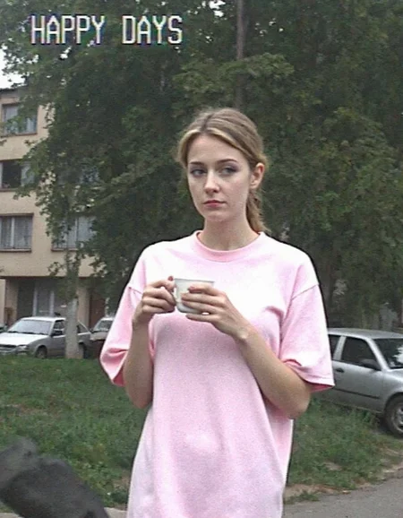 v8s, young woman, late 20s casually dressed in an oversized pink T-shirt, outdoors, her gaze directed to the side, sad expression, hold cup of coffee, her blonde hair loosely tied back. Her face partially shadowed, minimal makeup visible. she is standing on the street near soviet-era storey buildings, trees, parked cars near. Dull and overcast lighting. text overlay reading "HAPPY DAYS" in white pixelated font in top left corner. Image is heavily blurred, low resolution, grainy texture, overexposed in areas, amateurish quality, appears to be from vintage video footage or security camera, motion smeared details.