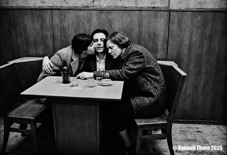 A black and white photograph of two women and a man sitting at a table in a dimly lit room. The woman on the left is kissing the man on the right, who is sitting in the middle of the image. Both women have short hair and are wearing tweed jackets. The man is wearing a suit and has a serious expression on his face. The table between them has a bottle of beer, a cup, and a saucer. The room has wooden paneling on the walls and a wooden floor. The image has a vintage feel <lora:_Flux\Anders_Petersen.safetensors:0.9:0.9>