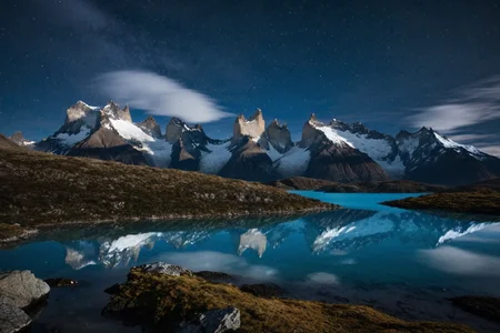 On the wind-blasted ridges of Torres del Paine in Chilean Patagonia, where jagged peaks tear through the clouds and icy lakes sit below. At moonlit night, (pale rock glow:1.2) traces the ridgeline, water lies still below, and stars flicker low.,