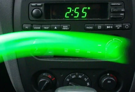 d1g1cam, Close-up image of a car audio system console, featuring a digital display reading 2:55 in bright green digits, surrounded by various control buttons for volume, bass, treble, and radio frequency adjustment. The buttons are arranged in a clean, organized manner, characteristic of late 90s or early 2000s car stereo designs. A vivid green, ethereal streak, possibly due to light reflection, arcs across the console creating a ghost-like effect. This unusual visual element adds a surreal touch to the otherwise standard car interior. The overall image quality suggests blurred details and an amateurish quality, possibly taken in motion or with an unsteady hand.,